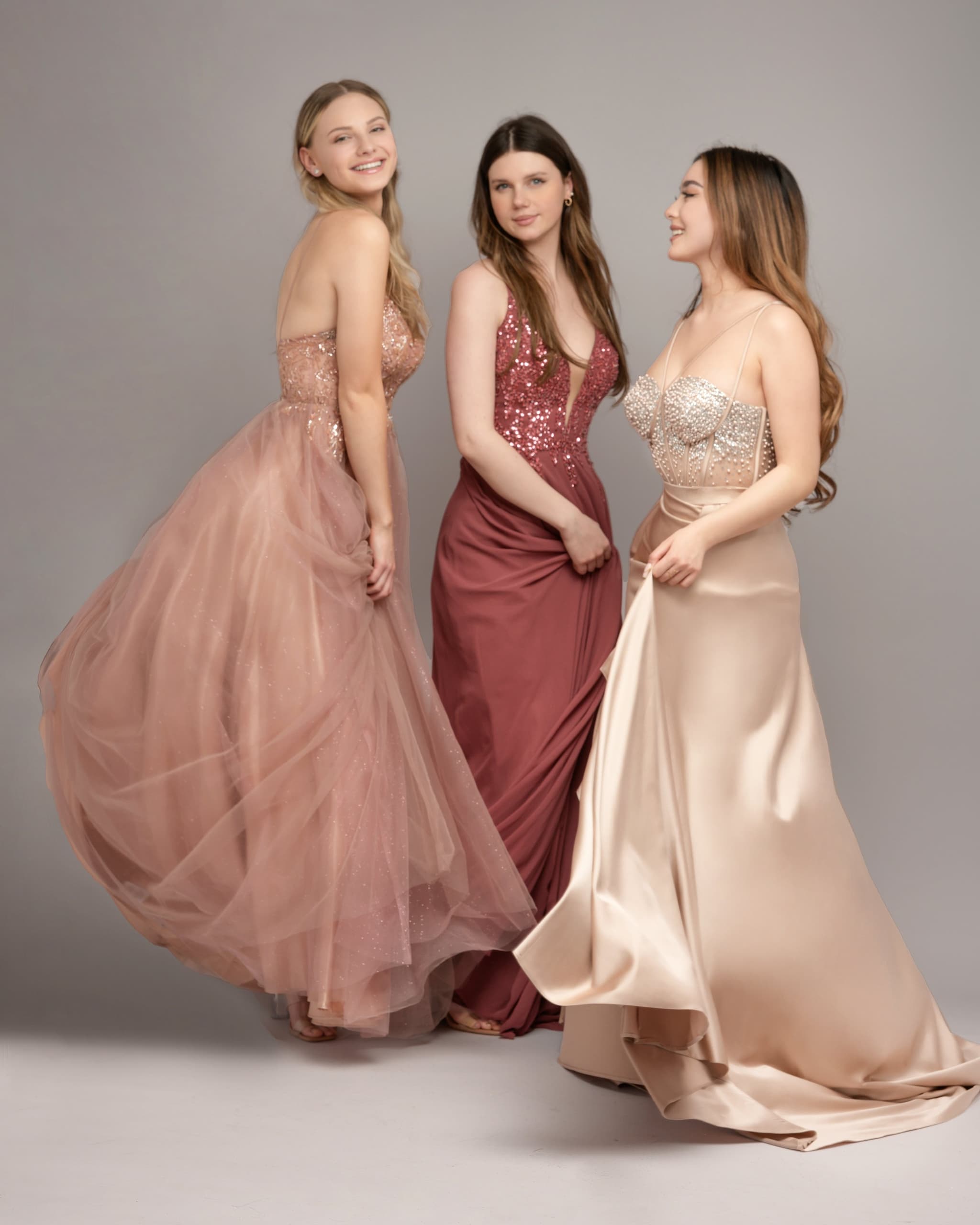 Drei junge Frauen in eleganten Abiballkleidern in Rosa, Burgund und Champagne mit Pailletten-Details, Gruppenfoto